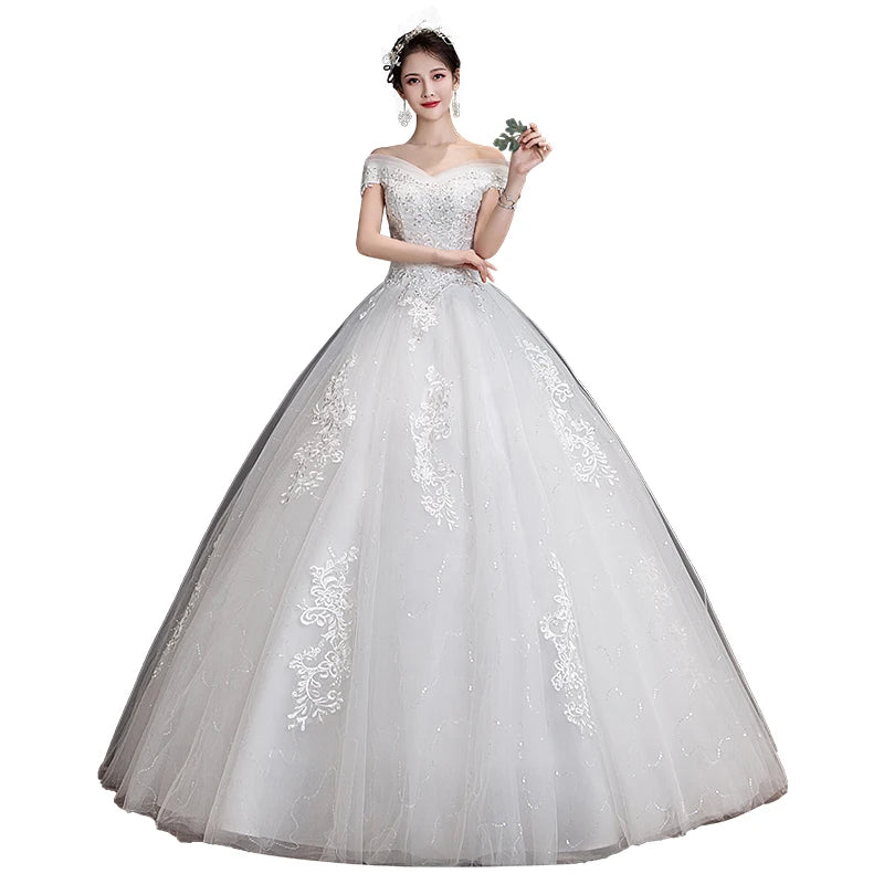 Woman in an elegant, off-shoulder white wedding gown adorned with lace patterns, holding a small flower. The dress features a full, flowing skirt and delicate detailing.