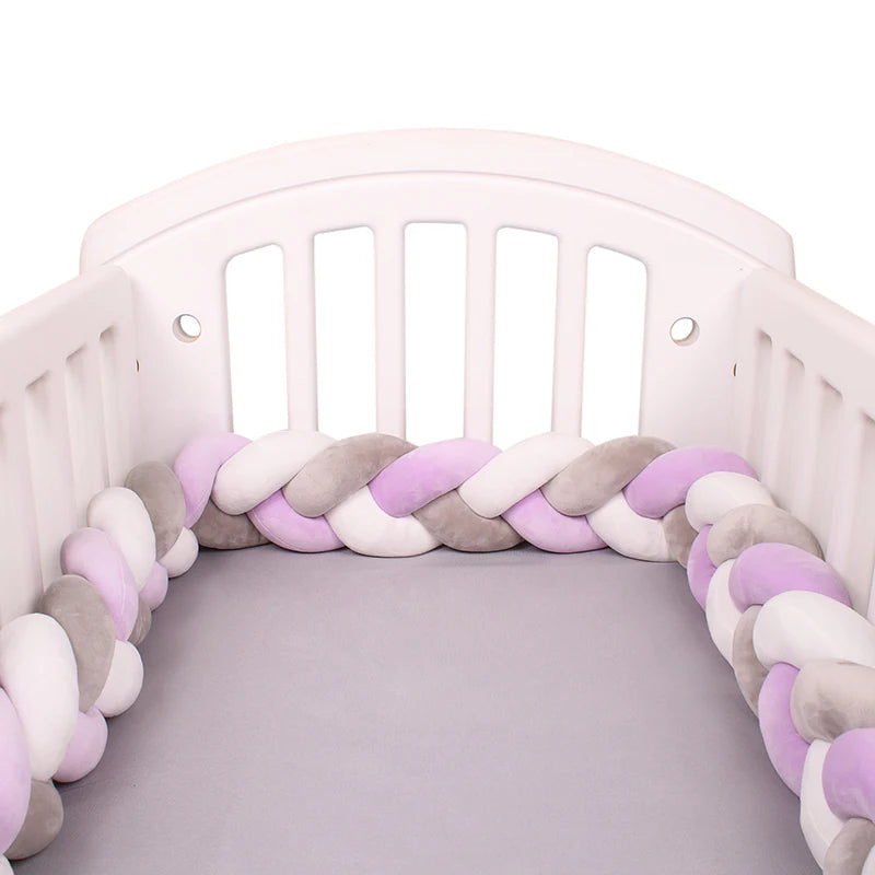 White crib with a braided bumper in pastel shades of pink, gray, and white, placed on a light gray mattress. The design adds a soft, cozy look to the crib's interior.