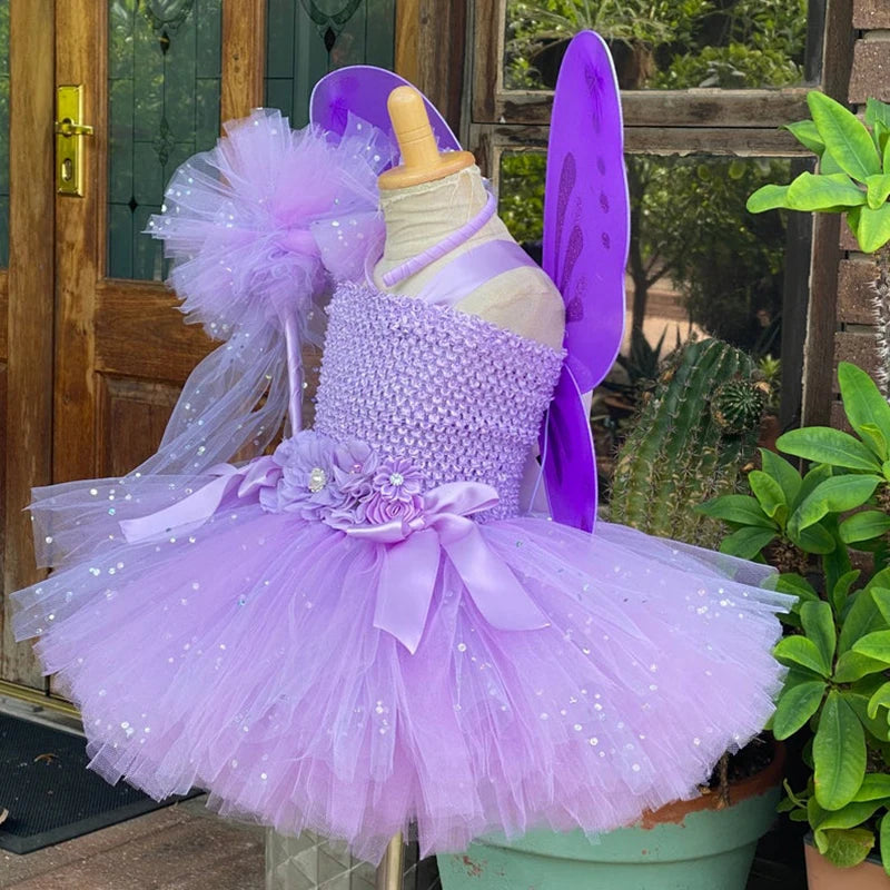 Lilac tutu dress with sequins and floral accents on a mannequin, featuring purple fairy wings. Set outside near a wooden door, surrounded by green potted plants.