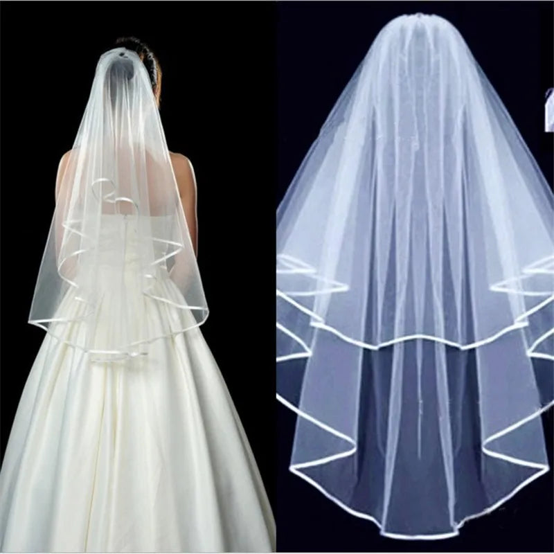A bride with a two-tiered white veil showcasing a satin-trimmed edge. The veil elegantly drapes over her back, enhancing the plain white gown beneath against a dark background.