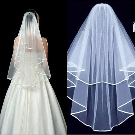 A bride with a two-tiered white veil showcasing a satin-trimmed edge. The veil elegantly drapes over her back, enhancing the plain white gown beneath against a dark background.