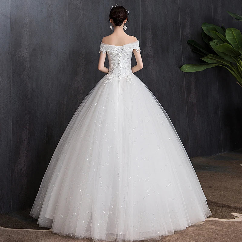 A woman in an off-shoulder, lace-embellished wedding gown with a full tulle skirt stands facing a dark textured wall, with a potted plant nearby.