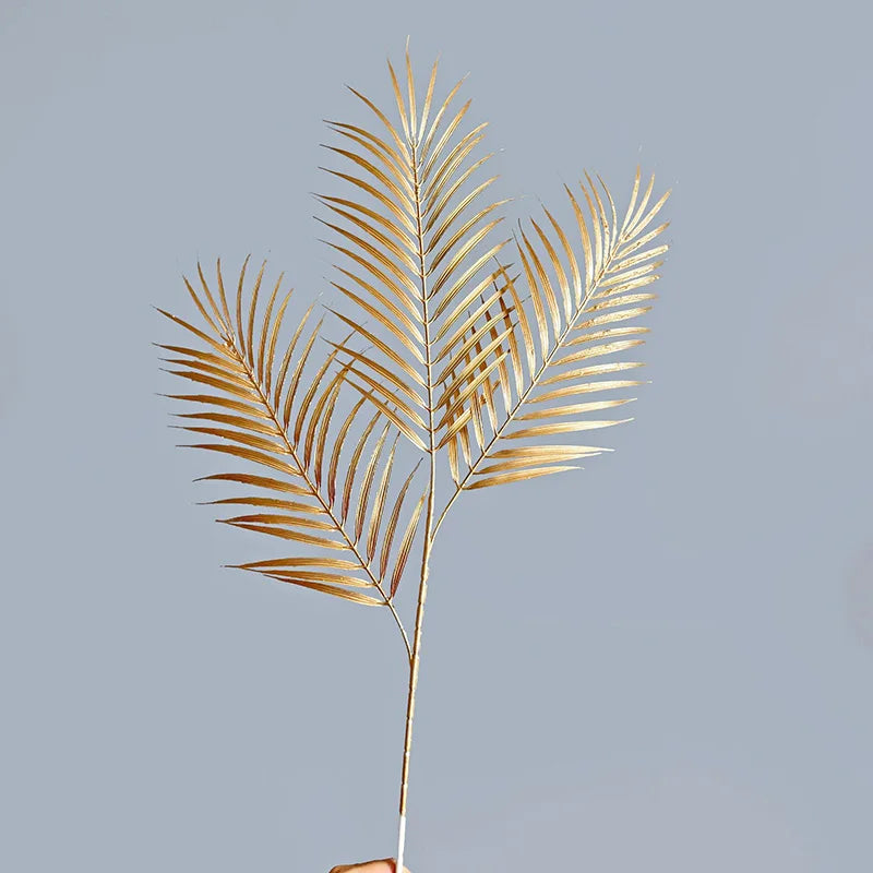Golden palm leaf with three fronds held against a plain gray background. The metallic shimmer highlights the intricate details of the leaf's structure.