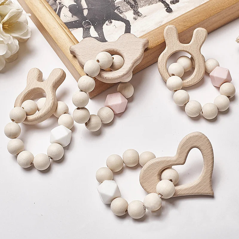 Wooden baby teethers with animal and heart shapes, featuring natural beads and hexagonal accents in soft pink and white tones, displayed on a table with a vintage photo in the background.