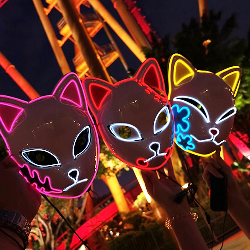 Three illuminated cat masks with neon outlines are held up in front of a brightly lit amusement park ride at night, showcasing pink, blue, and yellow designs.