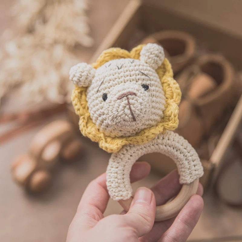Hand holding a crocheted lion-shaped baby rattle with a wooden ring. Background shows blurred wooden toys and baby shoes in a box.