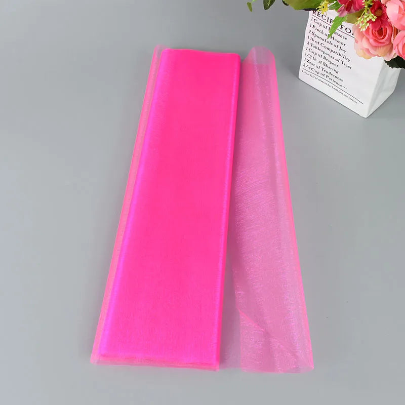 A bright pink organza fabric folded neatly on a gray surface. A white bag with a recipe and a bouquet of pink roses is visible in the background.