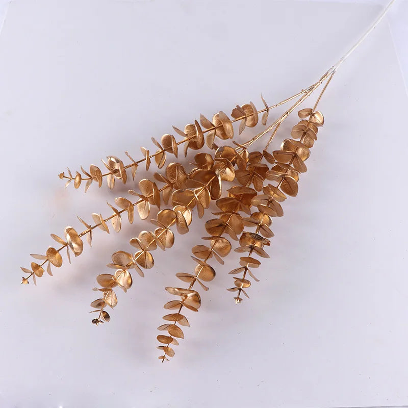 Gold-painted eucalyptus branches with multiple round leaves are arranged against a plain white background, creating a simple yet elegant display.