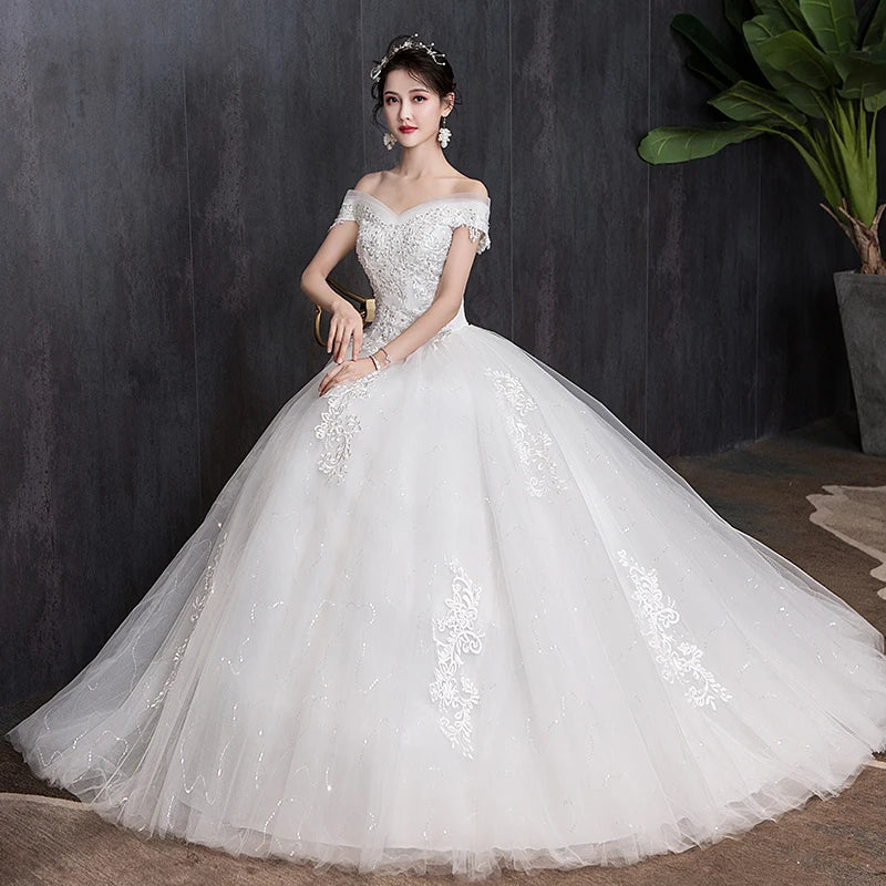 Bride in an elegant off-shoulder lace wedding gown with a full tulle skirt, seated indoors against a dark background, adorned with floral jewelry and surrounded by potted plants.
