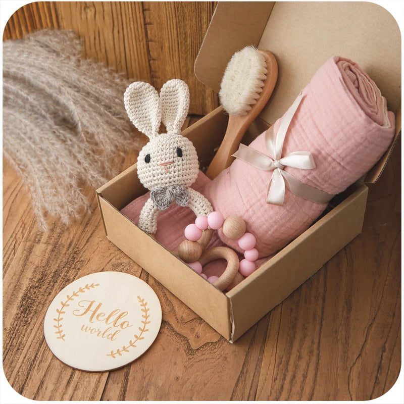 Gift box with a knitted bunny, pink swaddle, wooden brush, and a pink bead teether on a wooden surface. Nearby, a round wooden coaster reads "Hello world." Pampas grass decorates the background.