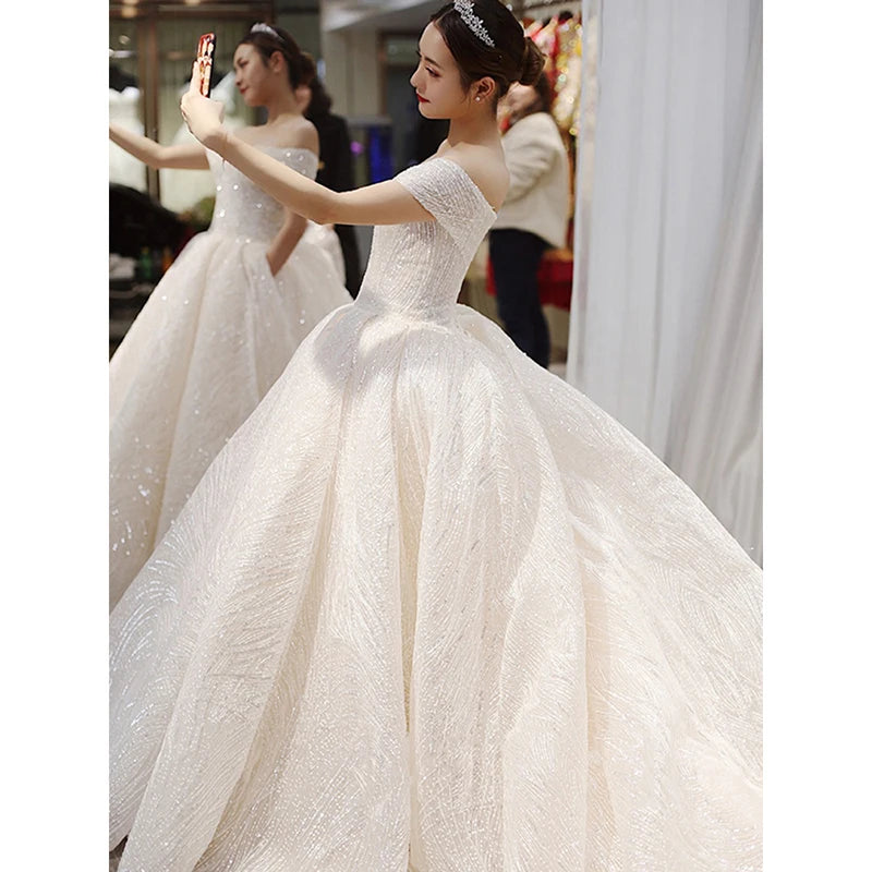 Woman in elegant off-shoulder wedding gown takes a selfie. The dress features a full, textured skirt and she's wearing a tiara, standing near a mirror reflecting her image.