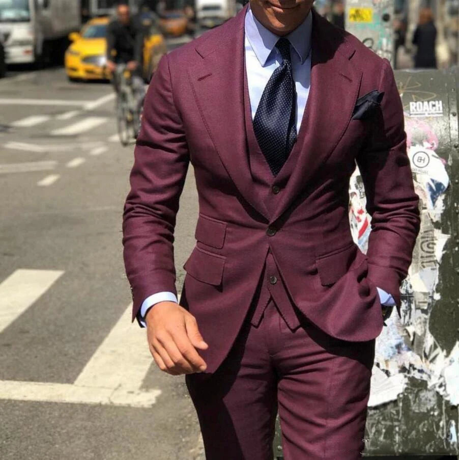Man in a fitted maroon suit with a navy tie and pocket square walks on a city street. Background includes a yellow taxi, cyclist, and a poster-covered wall.