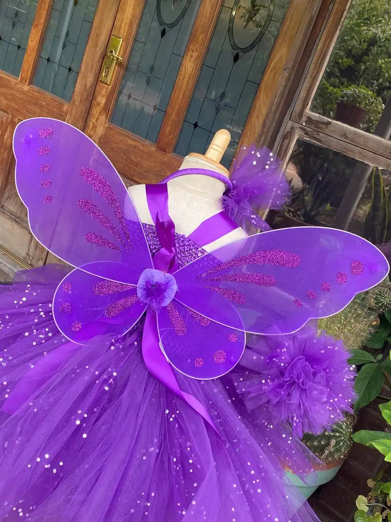 Purple fairy dress with sparkling wings and tulle skirt on a mannequin in front of a wooden door. The dress features glittering details and floral accents.
