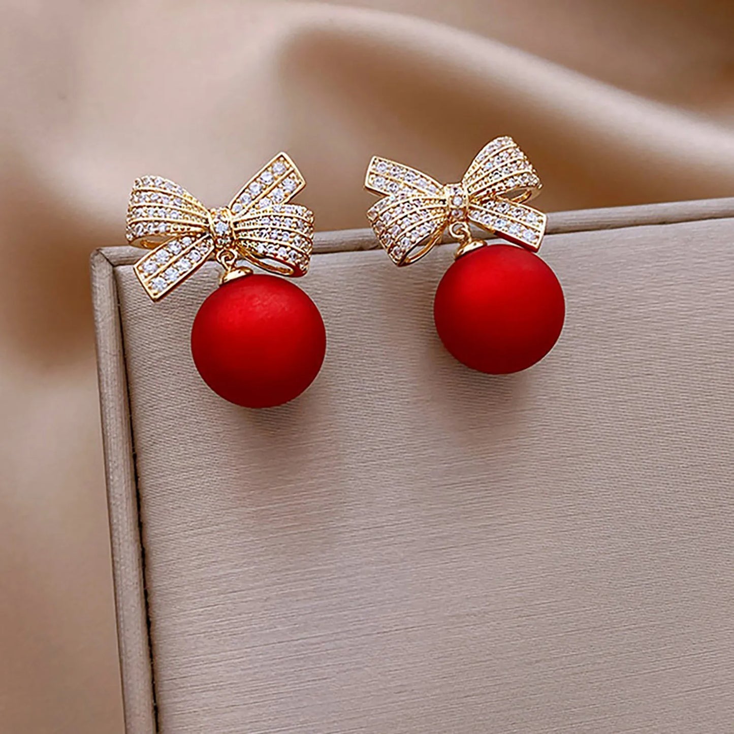 Elegant earrings with sparkling, gold bows studded with rhinestones and dangling matte red spheres, displayed on a soft beige fabric background.