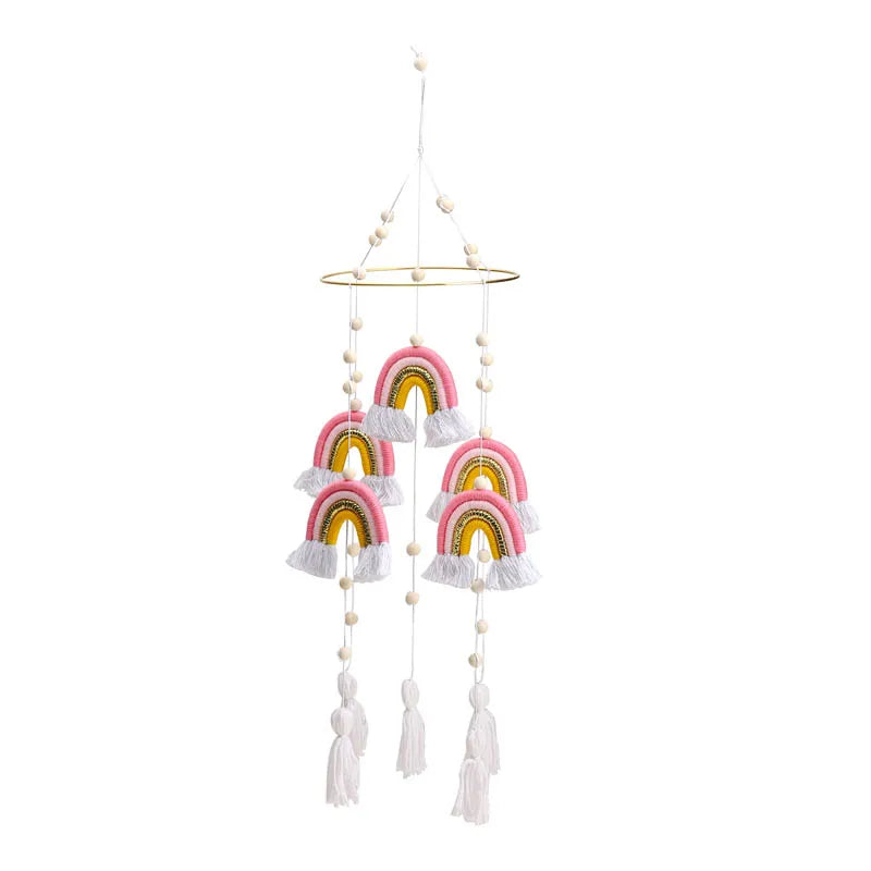 Hanging mobile with six yarn rainbows in pink, yellow, and white, featuring tassels and wooden beads. The rainbows are suspended from a circular gold ring.