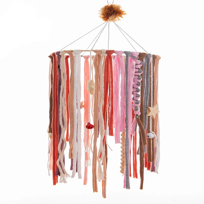 Hanging mobile with colorful fabric strips, beads, and wooden star shapes, topped with a fluffy pompom. The design is in warm pastel tones including pink, orange, and beige.