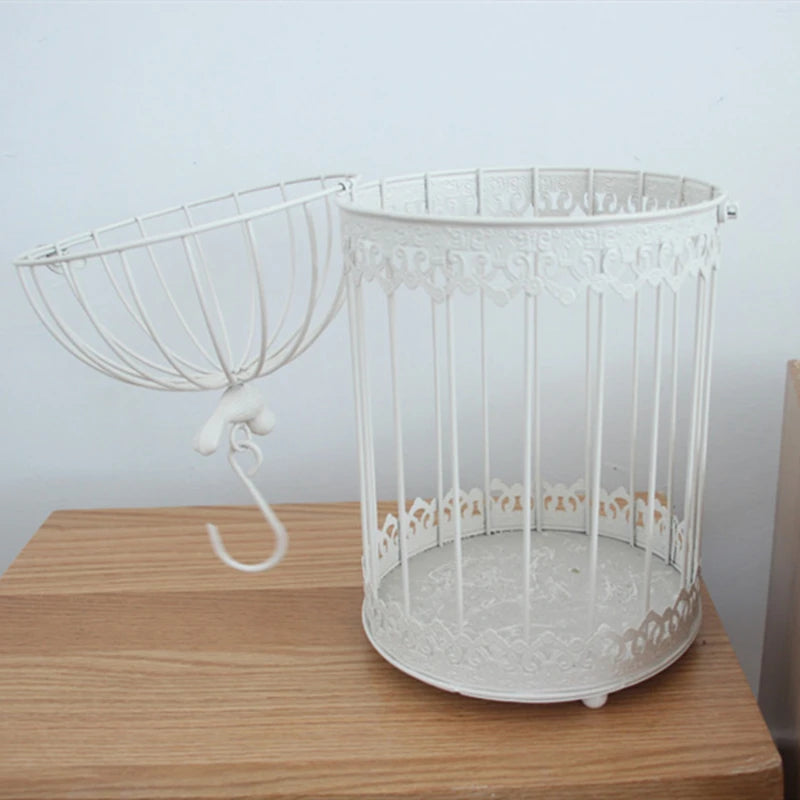 Ornate empty white metal birdcage with a decorative open top and hanging hook, placed on a wooden surface against a plain light background.