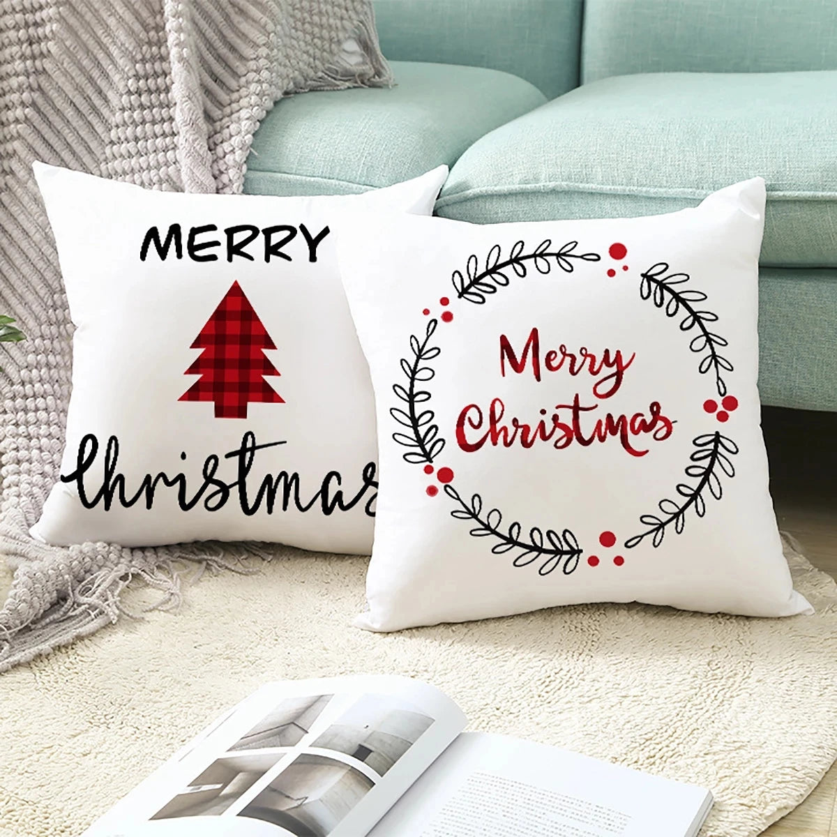 Two festive pillows on a beige rug and light blue sofa. One reads "Merry Christmas" with a red plaid tree; the other has a wreath design with red accents. An open book lies in front.