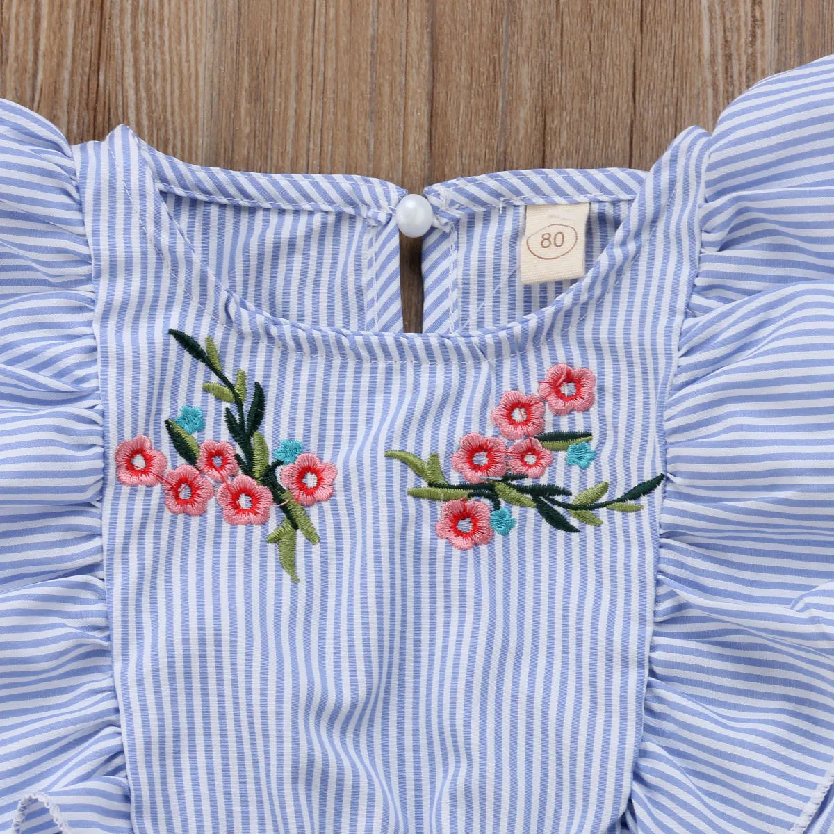 Blue and white striped garment with ruffled sleeves, featuring pink and green floral embroidery. A pearl button and tag marked "80" are visible at the back neck area.