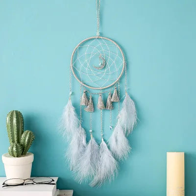 A dreamcatcher with a crescent moon hangs on a turquoise wall. It features silver tassels and light blue feathers. Nearby are a small cactus, a pair of glasses, and a candle.