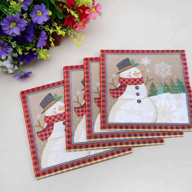Festive napkins featuring a snowman with a red plaid scarf and black hat, bordered in red plaid. Flowers in purple, pink, and yellow accompany the napkins on a light surface.