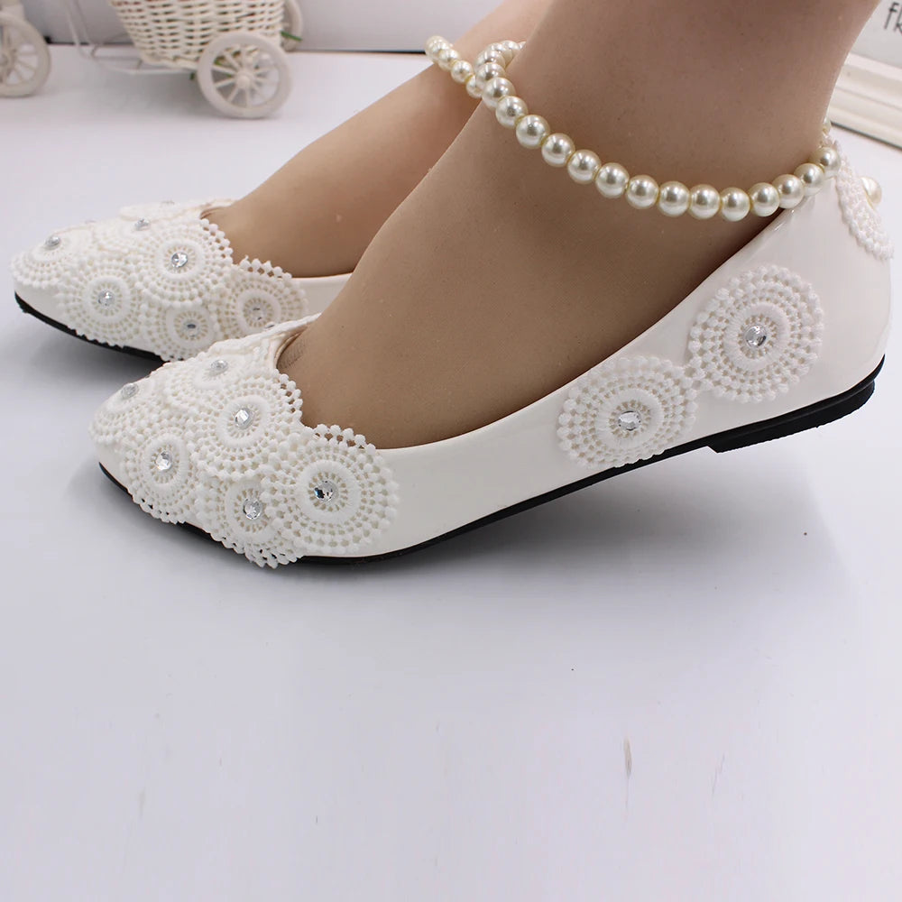 White flats with decorative circular beading and rhinestones, paired with a pearl ankle bracelet. Background shows a small decorative white cart on a white surface.