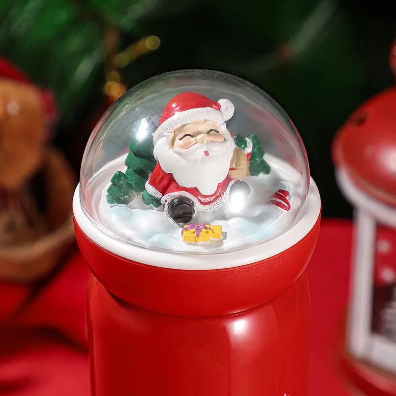 Festive snow globe atop a red container features a jolly Santa in a red suit, sitting on snow with a present and a small tree, set against a blurred, colorful holiday background.