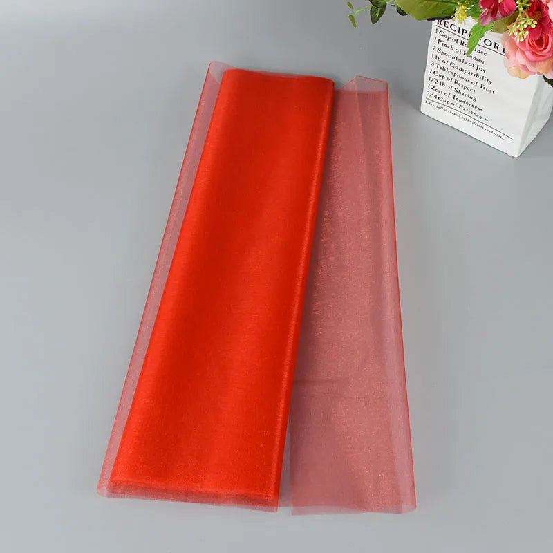 A red shimmery tulle roll unfurled on a gray surface, with a bouquet of pink flowers and a decorative box nearby.