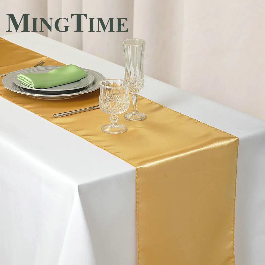Elegant table setting with a gold satin runner over a white tablecloth. Includes a green napkin on a plate, cutlery, and crystal glassware. MingTime logo in the background.