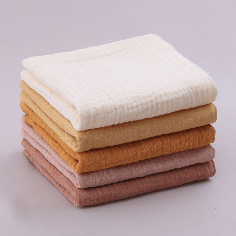 A neatly stacked pile of six folded gauze towels in earthy tones, including off-white, mustard, orange, light pink, dusty pink, and brown, against a light gray background.