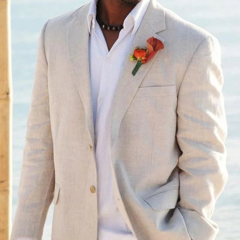 Man in a light beige suit with a white shirt and an orange boutonniere, standing against a blurred outdoor background, possibly near the water.