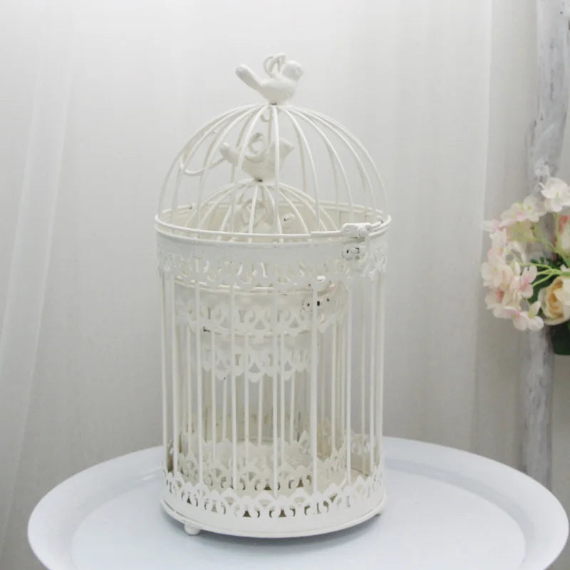 A decorative white metal birdcage with intricate patterns, topped with bird figurines, placed on a round white table. A bouquet of pastel flowers is softly blurred in the background.