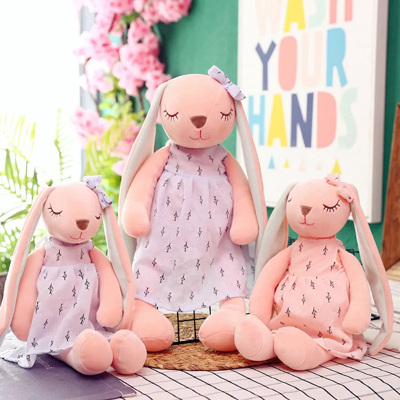 Three plush bunny toys with closed eyes in pastel dresses and bows sit on a table. A sign in the background reads "Wash Your Hands," surrounded by pink flowers and greenery.