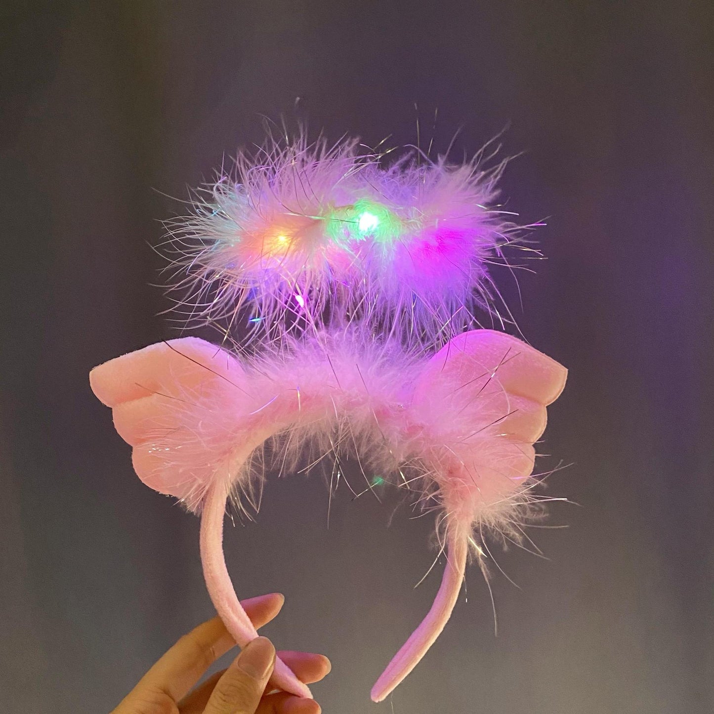 A pink, fluffy headband with angel wings and glowing multicolored LED lights is held in a hand against a dark background.
