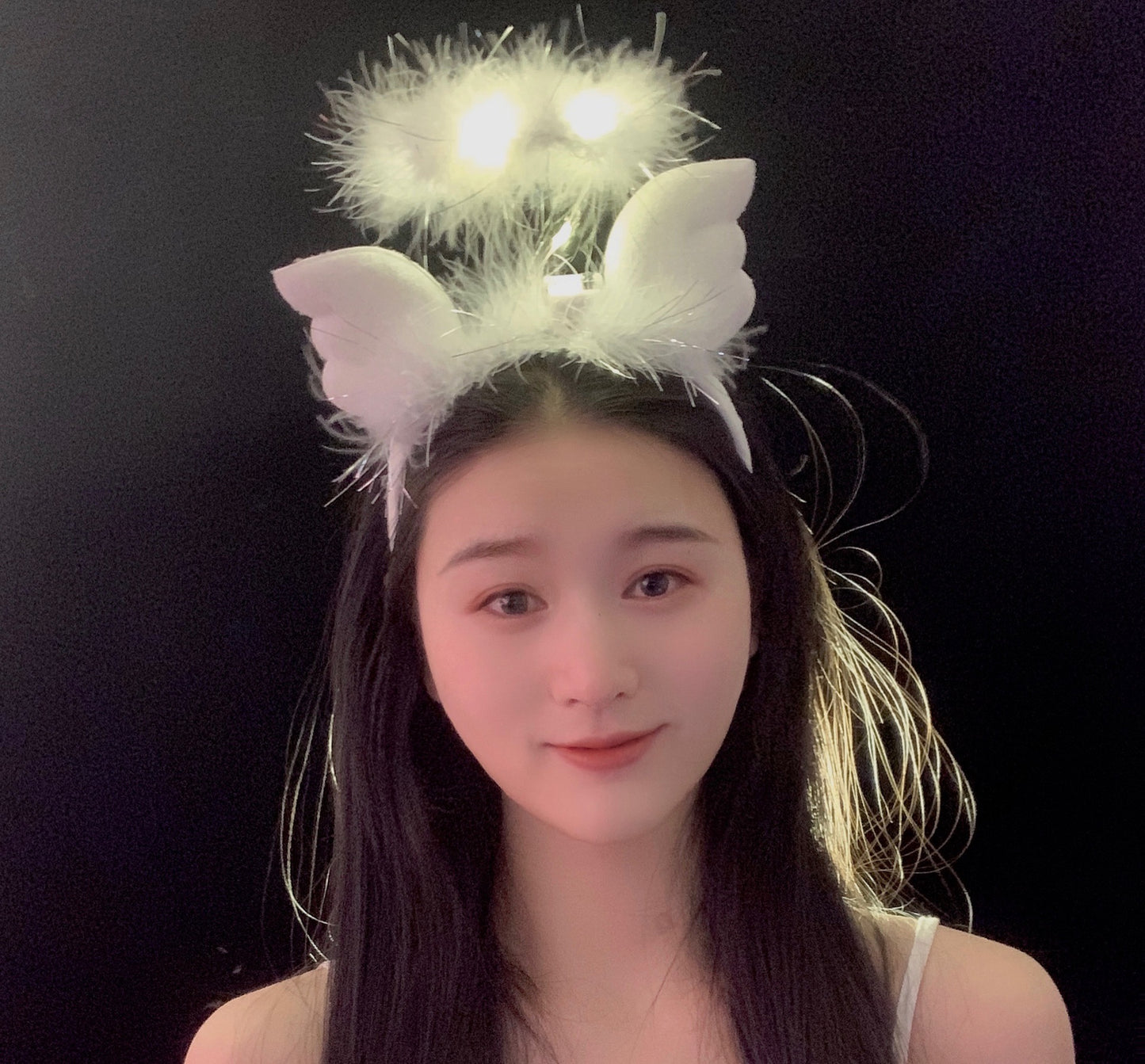 A person with long black hair smiling, wearing a white fluffy headband with glowing lights and attached wings, set against a black background.