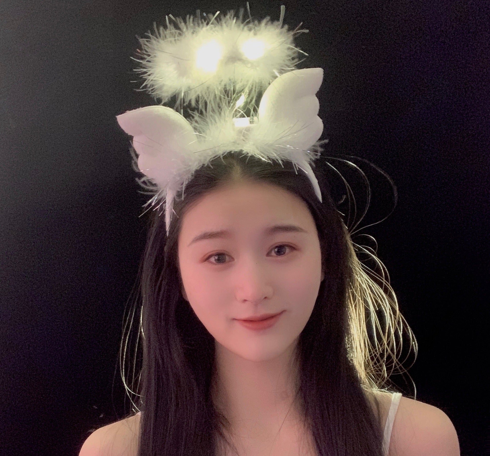 A person with long black hair smiling, wearing a white fluffy headband with glowing lights and attached wings, set against a black background.