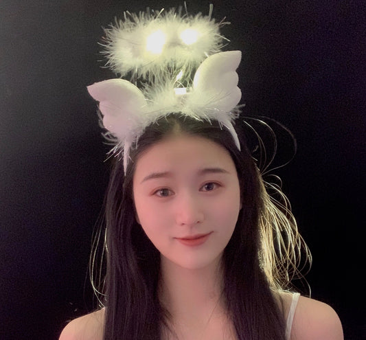 A person with long black hair smiling, wearing a white fluffy headband with glowing lights and attached wings, set against a black background.