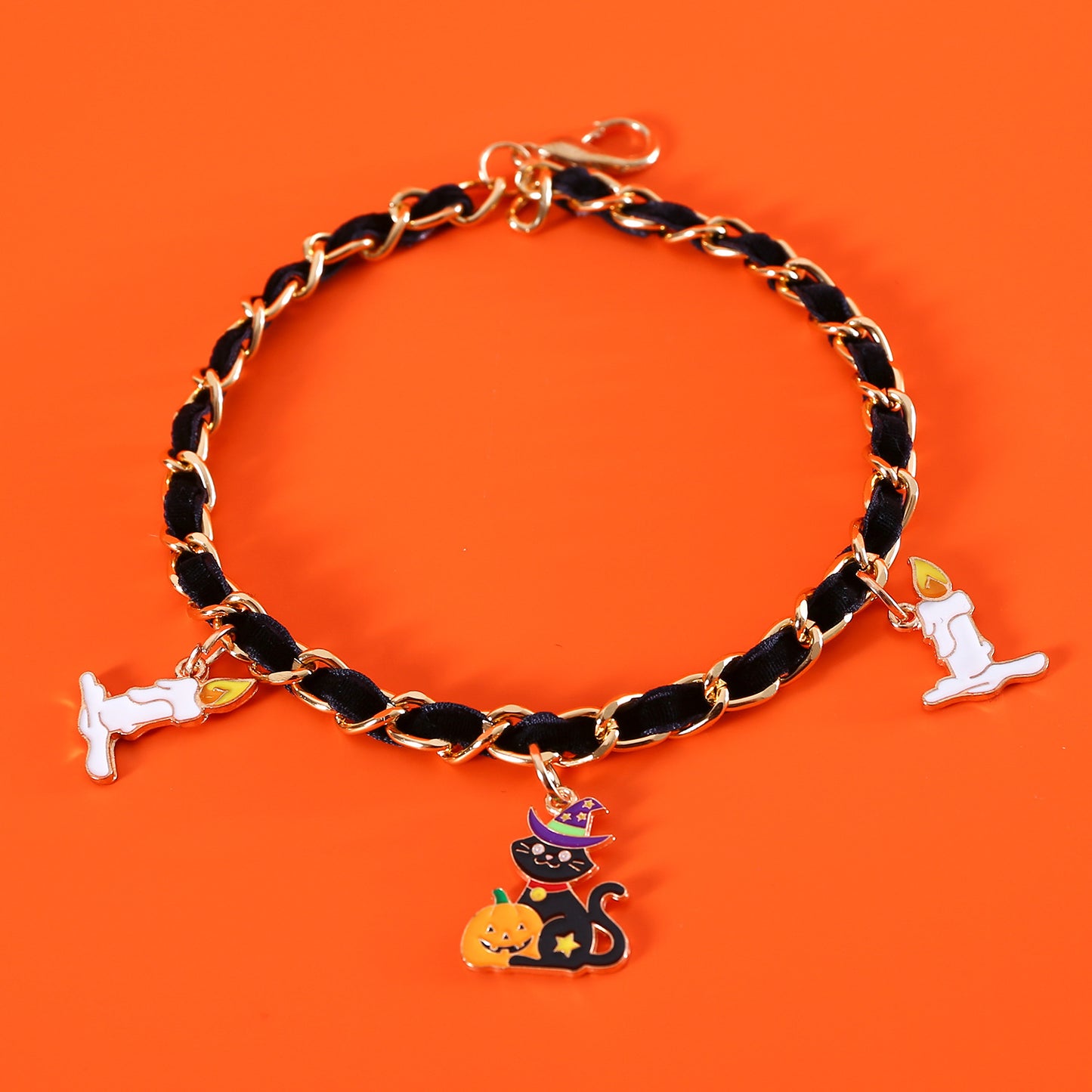 A black and gold chain bracelet with Halloween charms, including a black cat with a witch hat and pumpkin, flanked by two white candles on an orange background.