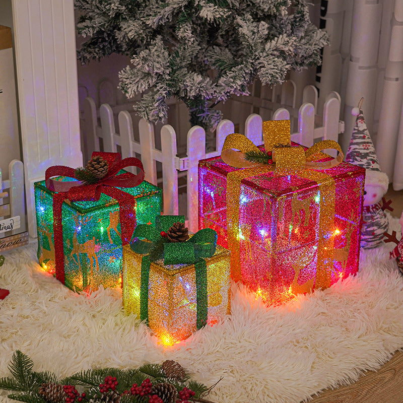 Three glittery gift boxes with bows and pinecones are illuminated by multicolored lights under a snowy Christmas tree beside a white picket fence on a fluffy white rug.