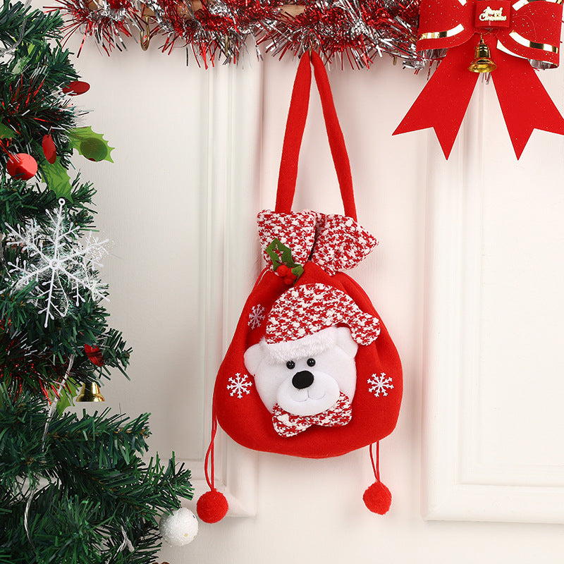 Red Christmas gift bag with a bear face, snowflake decorations, and a plaid hat, hanging on a wall near a decorated tree and red ribbon bow with a small bell.