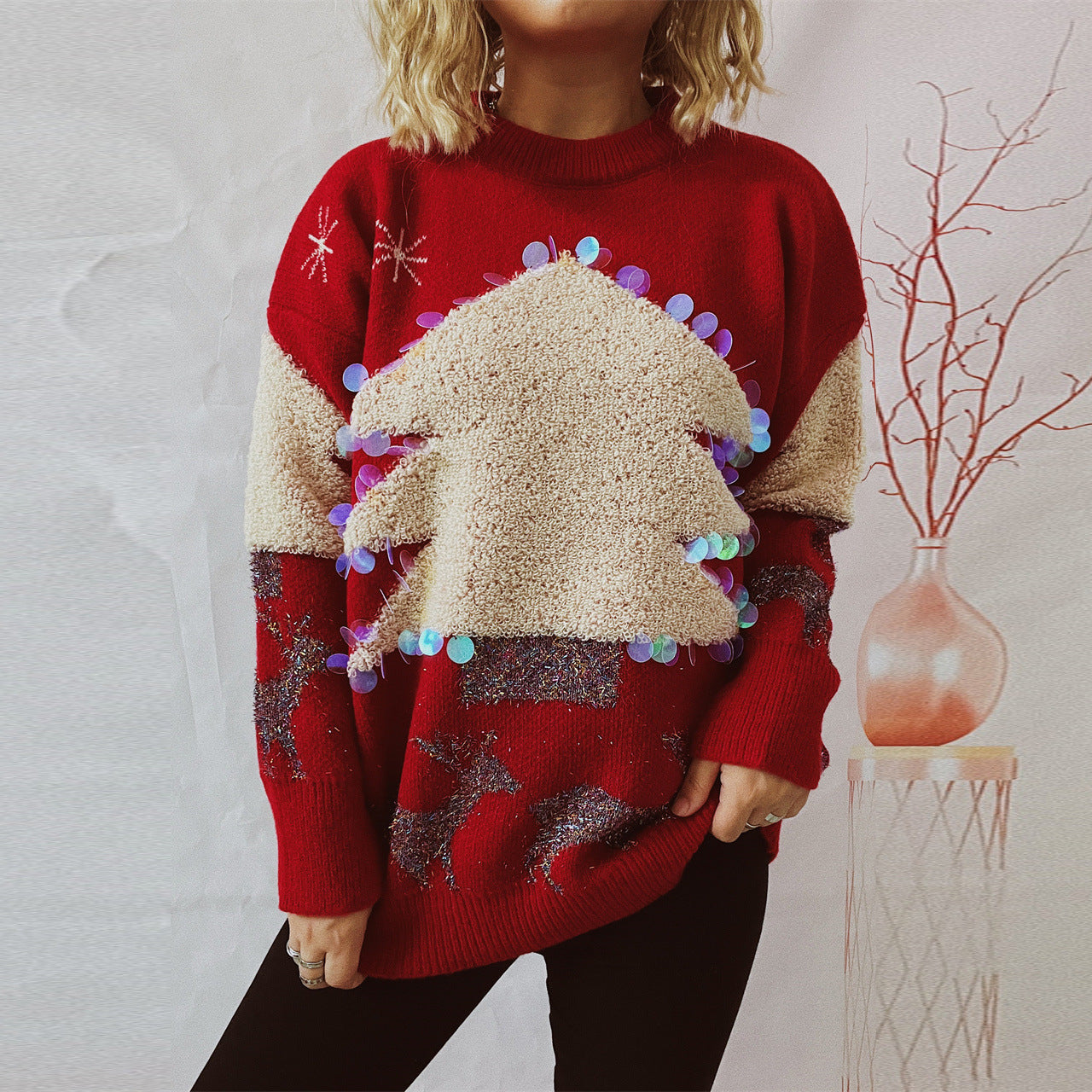 Woman wearing a red holiday sweater with a textured, sequin-adorned Christmas tree and sparkly reindeer design. She stands against a neutral background near a vase with branches.