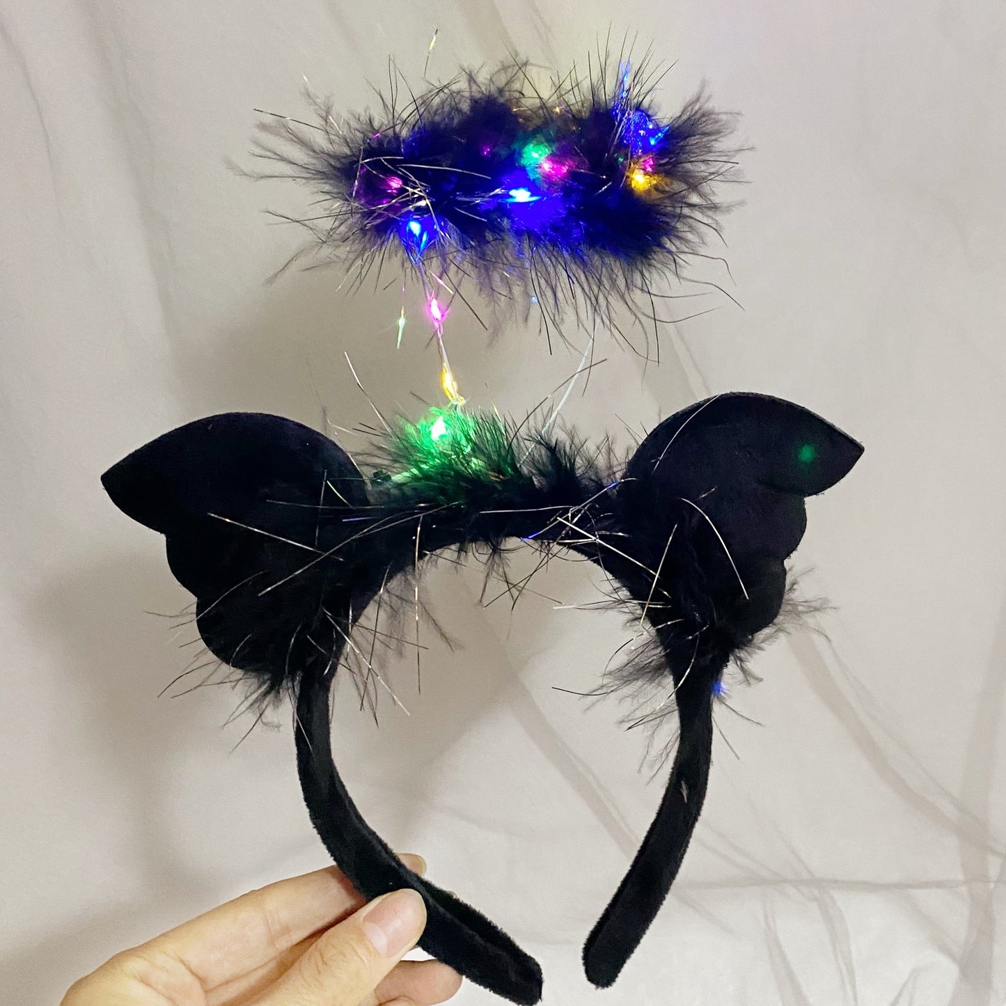 Black cat ear headband with feather trim and a halo made of black feathers adorned with colorful lights, held in hand against a plain background.