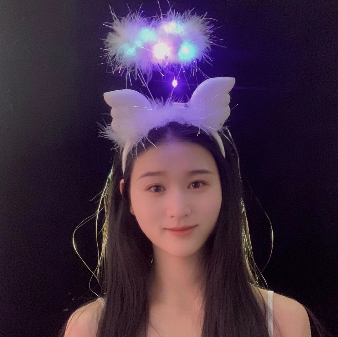 Young woman with long dark hair, wearing a white fluffy headband with glowing lights, resembling a halo and cat ears, against a black background.