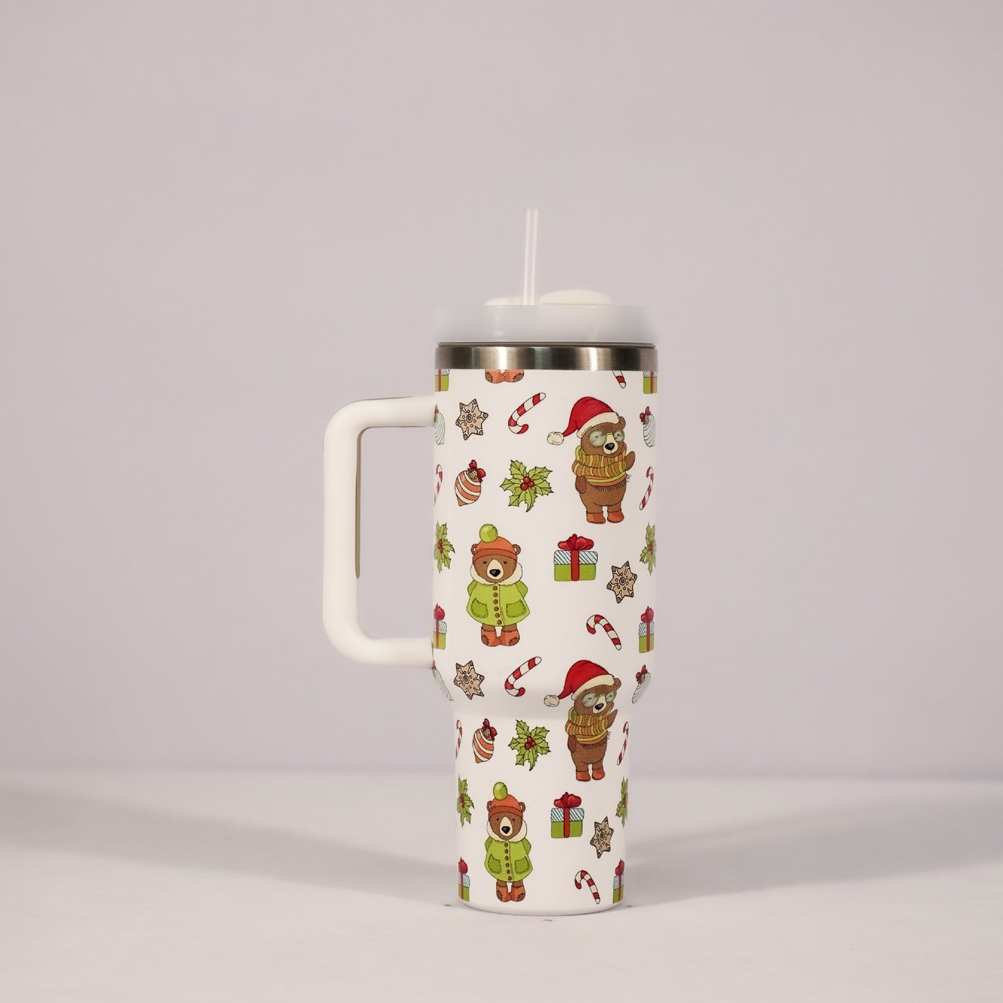 White travel mug with handle, decorated with festive bears in winter clothes, candy canes, holly, and gifts. Silver rim and white lid with a straw. Pale background.