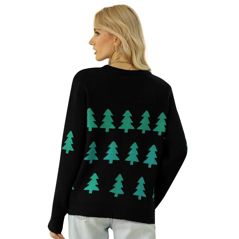 Woman with blonde hair wearing a black sweater featuring green tree patterns, turning slightly to show the back. She's paired it with light-colored jeans and gold earrings.