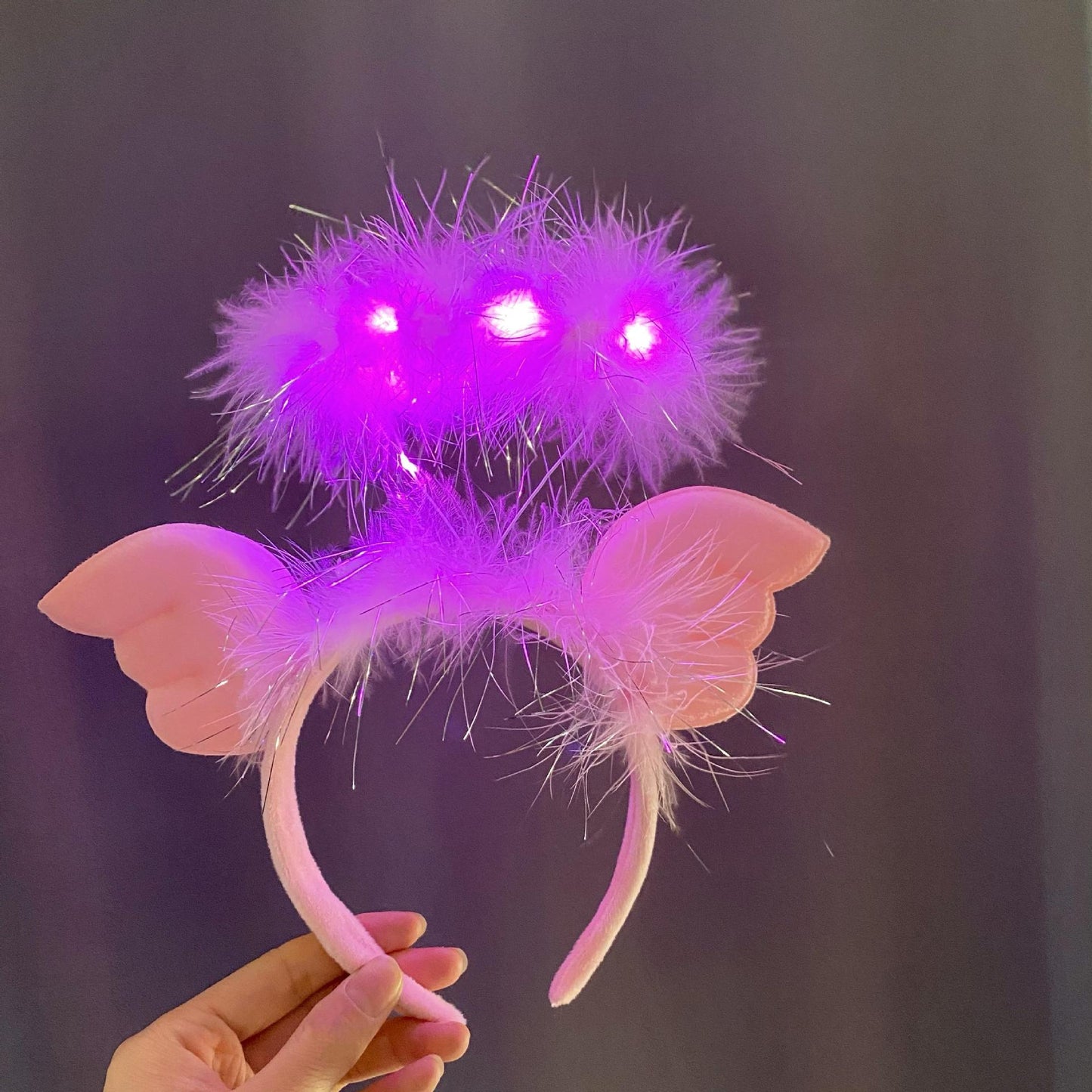 A hand holds a pink headband featuring fluffy feathers with glowing purple lights, adorned with pink, wing-like shapes on the sides, against a gray background.