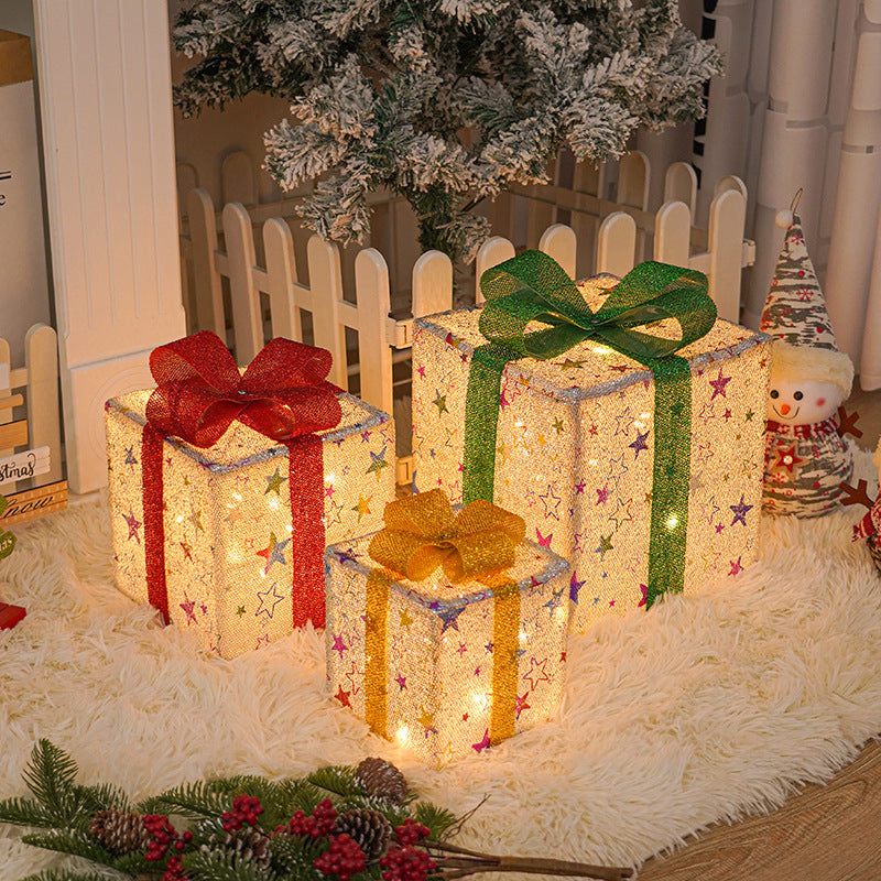 Illuminated Christmas gift boxes with colorful bows sit on a shag rug near a festive tree and picket fence, surrounded by holiday decor and a snowman figurine.