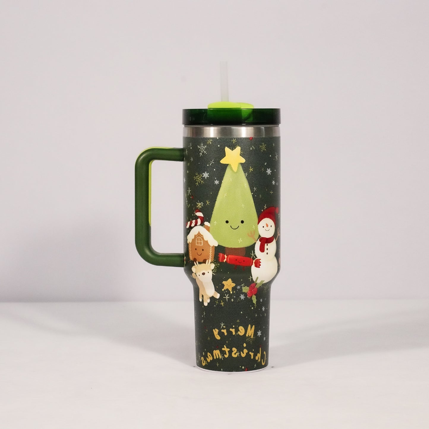 Festive travel mug with a smiling green tree, snowman, gingerbread house, and reindeer on a dark background. Features a green lid with a straw and "Merry Christmas" text.