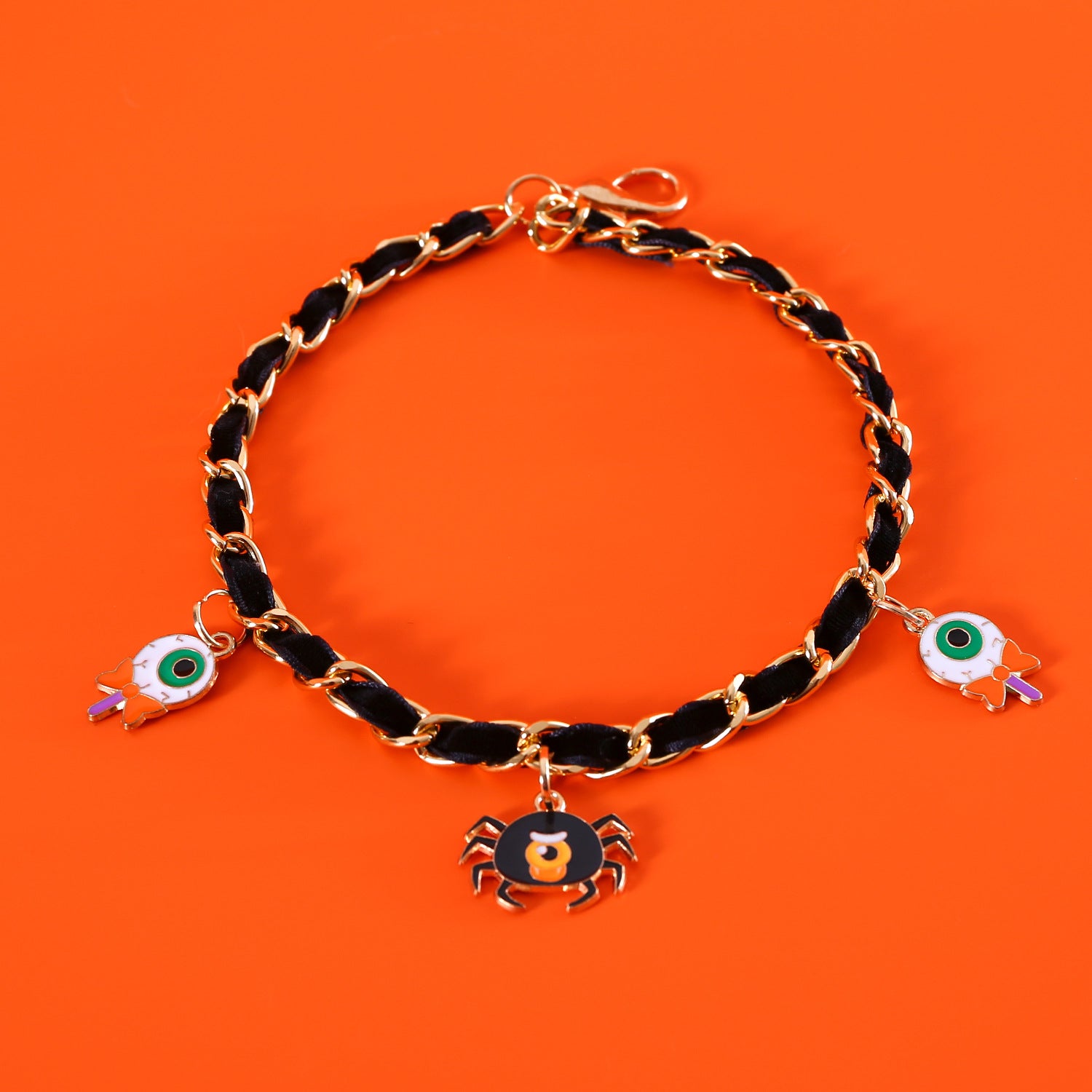 A gold chain bracelet with black woven fabric featuring Halloween charms: a spider and two eyeballs against an orange background.