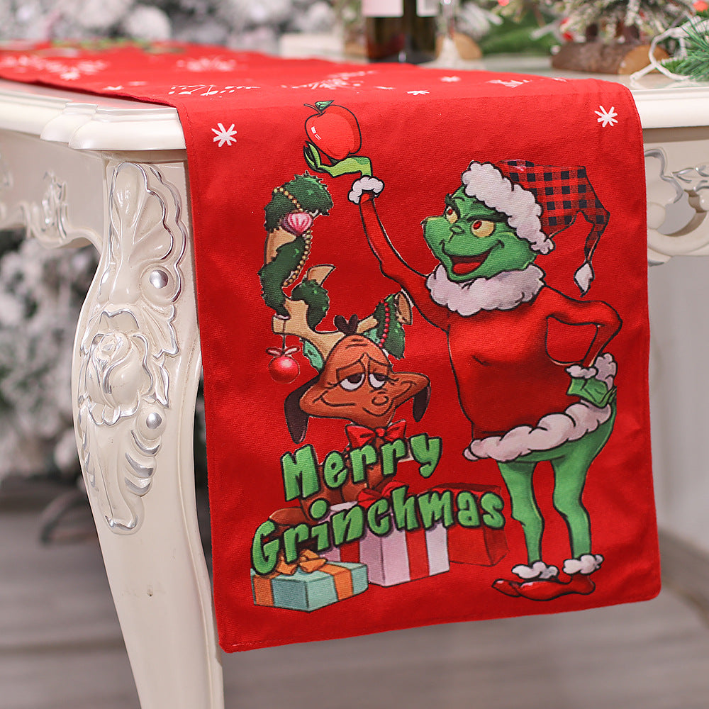 Festive table runner featuring a green, mischievous character in a Santa outfit, holding an ornament alongside a reindeer with gifts. The text reads "Merry Grinchmas".